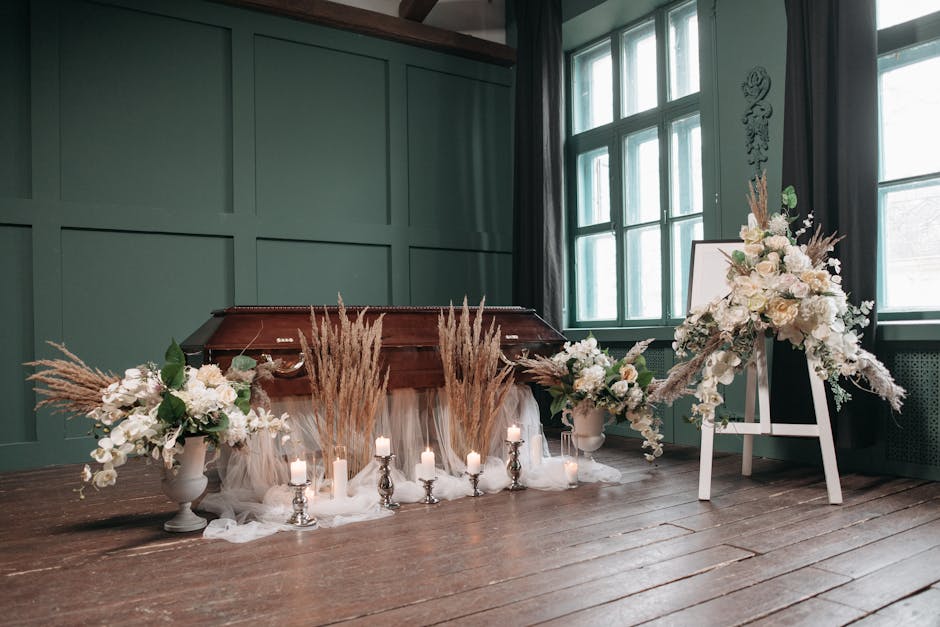 A floral arrangement featuring a large, elegant bouquet with white roses, lilies, and orchids, complemented by beige pampas grass and green foliage, displayed on a white easel and a pedestal vase in a