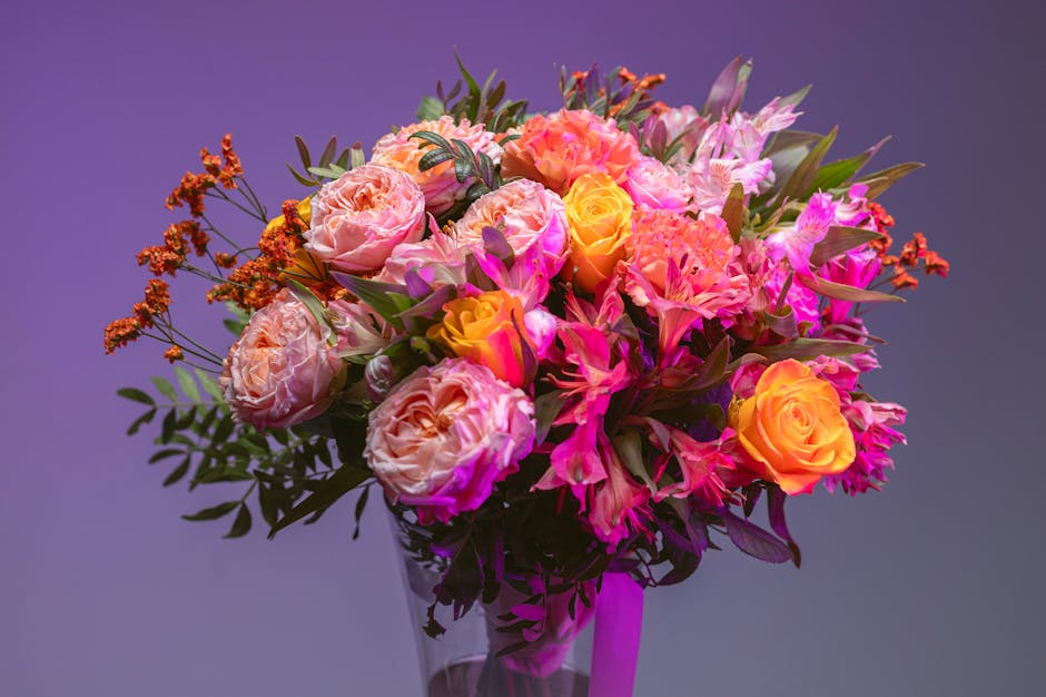 A vibrant bouquet of fresh flowers arranged in a clear glass vase, featuring large peach and pink roses, complemented by delicate pink lilies and smaller orange blossoms. The bouquet is lush with gree