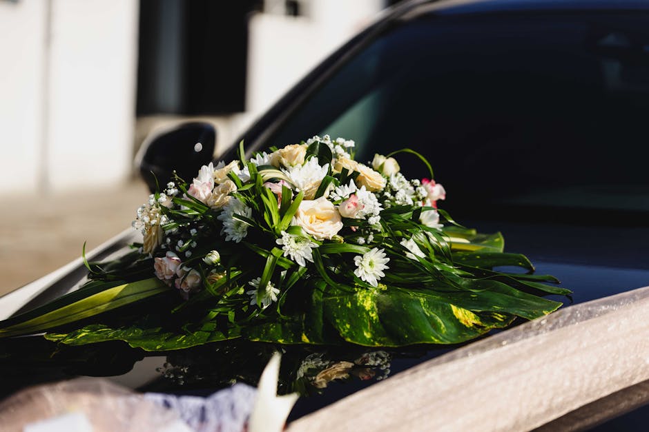 A floral arrangement featuring white and pale pink roses, white daisies, and small white filler flowers, arranged with lush green leaves and stems. The bouquet is placed on the hood of a black car, wr