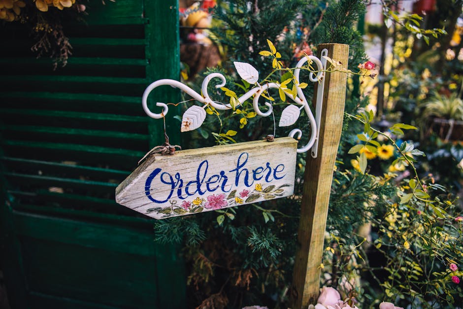 A rustic wooden sign with the word 'Order here' painted in blue cursive, hanging from a decorative white metal bracket attached to a vertical wooden post. The sign is adorned with small, delicate flor