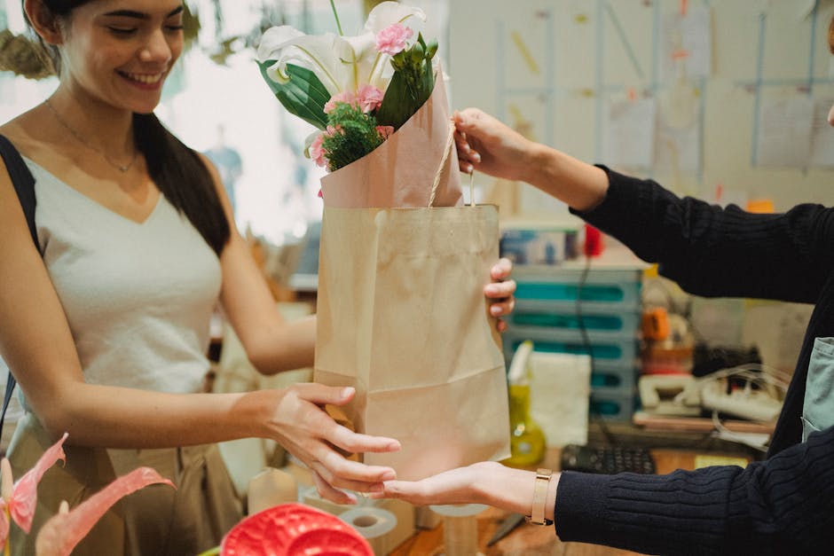 A woman with long dark hair, wearing a sleeveless white top, smiling as she receives a flower bouquet from another person in a floral shop. The bouquet features pink and white flowers, including lilie