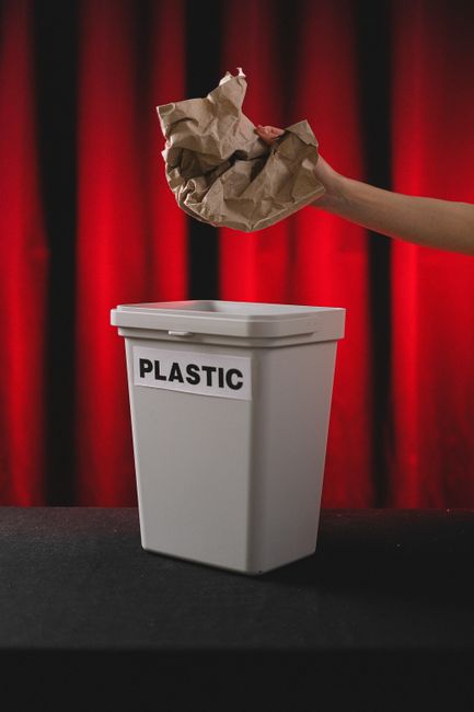 A person's hand is seen placing crumpled brown paper into a white plastic bin labeled 'PLASTIC' against a backdrop of red curtains. The scene is set on a black surface, and the focus is on the action