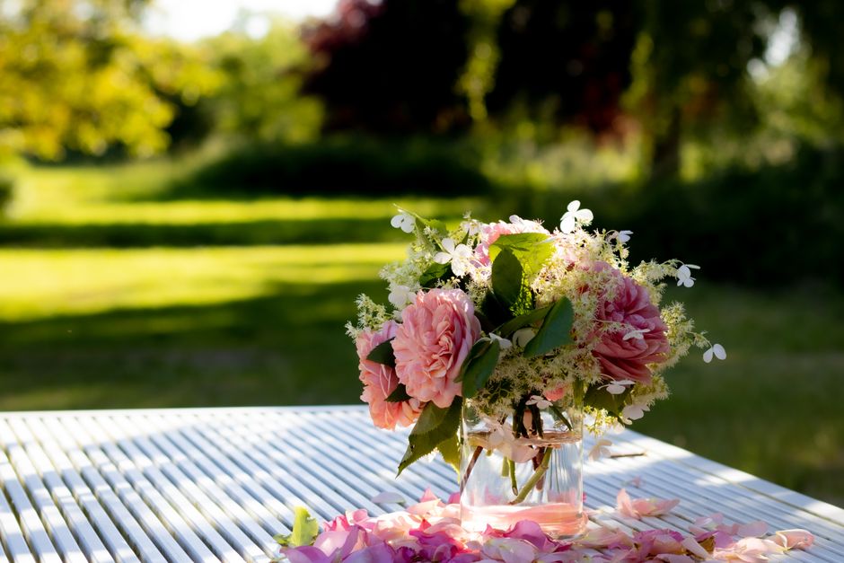 A delicate floral arrangement featuring large, soft pink peonies surrounded by small white flowers and green leaves in a clear glass vase. The bouquet is placed on a white slatted outdoor table, with
