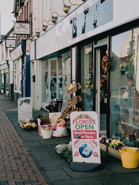 Outdoor view of a flower shop with large glass windows and an open sign, displaying an array of fresh flower arrangements and bouquets on the sidewalk. The floral displays include vibrant yellow, whit