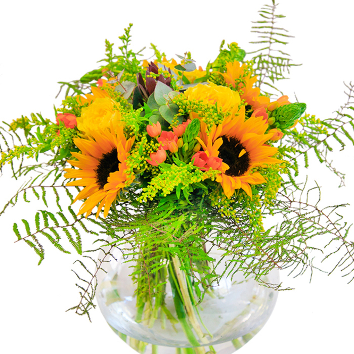 This golden-hued bouquet, arranged by Wallington Florist for same-day delivery across Wallington and nearby Carshalton, radiates an inviting, sunlit cheer reminiscent of afternoons in Beddington Park. Tall, bold sunflowers anchor the arrangement, each bloom's yellow-orange petals unfurling around dark, textured centers-like snapshots of the summer sun warming the local Green. Surrounding them, yellow garden roses spiral open in layers of silken petals, each one contributing a classic beauty and soft fragrance that whispers of local garden walks. Scarlet Hypericum berries nestle amongst the greens, their glossy surfaces catching the light, infusing the bouquet with playful energy and vivid colour-echoes of community gatherings on London Road. The scene is softened by a cascade of feathery fern fronds and airy, light green spirals of filler; delicate tufts of goldenrod speckle the arrangement with miniature yellow flowers, and eucalyptus leaves add a hint of blue-green coolness. All are gathered within a round, clear glass vase, the stems neatly displayed, their freshness easily seen and enjoyed by recipients throughout Wallington. This piece feels generously alive-every bloom, leaf, and berry thoughtfully placed to create texture and movement. It is an ideal choice for marking a special occasion, offering thanks, or simply brightening a space with the joyful abundance and locally grown charm that Wallington Florist is known for.